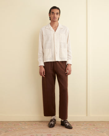 A person stands against a light-colored wall, wearing the Geo Lace Overshirt in Ecru with an intricate 1950s machine lace pattern and brown pants. The boxy fit of the overshirt adds a modern touch. They have dark hair and are standing on a patterned floor.