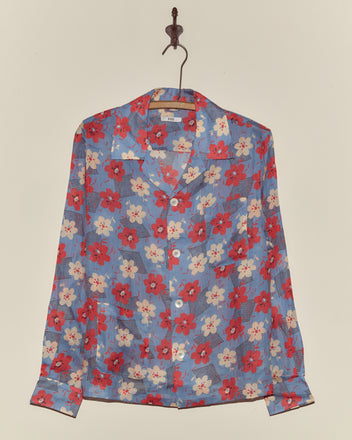 The Geo Poppy Long Sleeve Shirt, in cornflower blue with a red and white floral print inspired by 1940s style, hangs on a hanger against a beige wall.