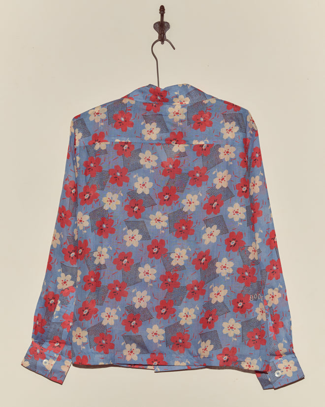 The Geo Poppy Long Sleeve Shirt, inspired by the 1940s, features a red and white floral print on a cornflower blue rayon background and hangs on a hook.