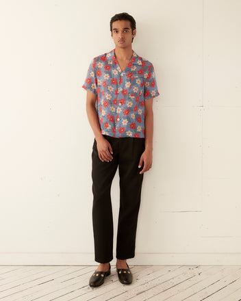 A person stands against a light-colored wall, wearing a blue Geo Poppy Short Sleeve Shirt with a floral design, black pants, and black shoes.