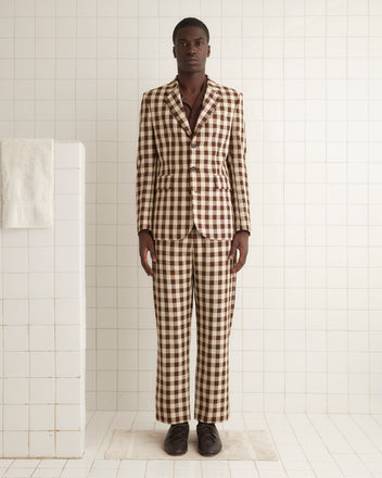 A person stands against a light-colored wall, adorned in a Gilbert Check Single-Breasted Suit Jacket made from deadstock fabric, featuring a brown and white checkered pattern reminiscent of the 1950s. Their outfit is completed with matching trousers and brown shoes.