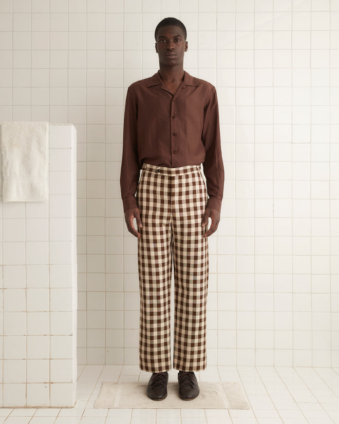 A person stands indoors against a plain wall, wearing a brown button-up shirt and the Gilbert Check Trousers, which feature 1950s-inspired checkered patterns and side-buckle waist adjusters. They have their hands in their pockets and are sporting dark lace-up shoes.