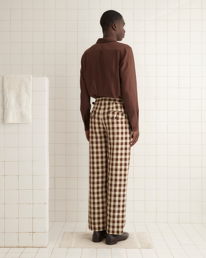 A person stands indoors against a plain wall, wearing a brown button-up shirt and the Gilbert Check Trousers, which feature 1950s-inspired checkered patterns and side-buckle waist adjusters. They have their hands in their pockets and are sporting dark lace-up shoes.