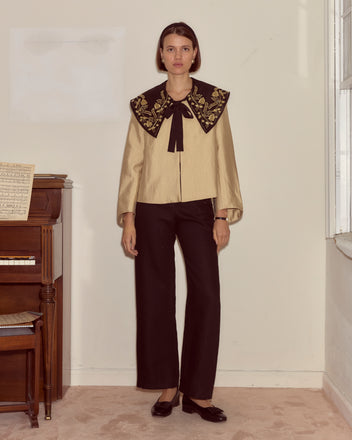 A woman stands indoors by a piano, wearing black pants and the Gilt Jacket, which features a gold blouse design with an embroidered collar inspired by Ottoman Empire gold bullion embroidery.