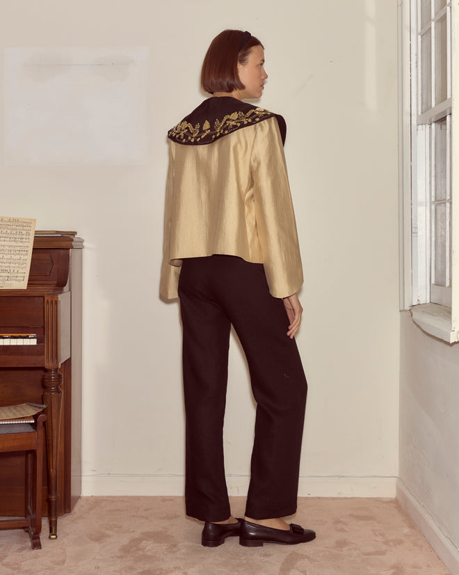 Indoors by a piano, a woman wears the Gilt Jacket—featuring a floral collar and ornate gold detailing inspired by Ottoman-era ceremonial towels—paired with black pants.