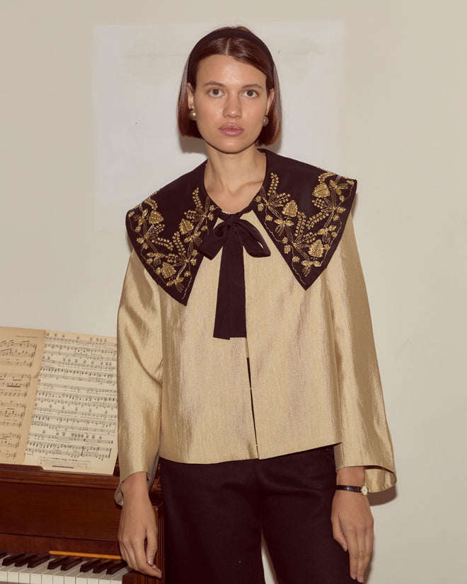 A woman wearing the Gilt Jacket—featuring gold bullion embroidery and a black embroidered collar—stands in front of a piano with sheet music.