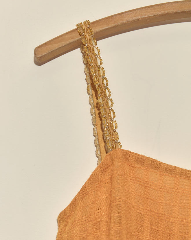 Close-up of the Gilt Rosetta Tank - S, a yellow textured dress with lace straps and vintage beaded trim, hanging on a wooden hanger against a light background.