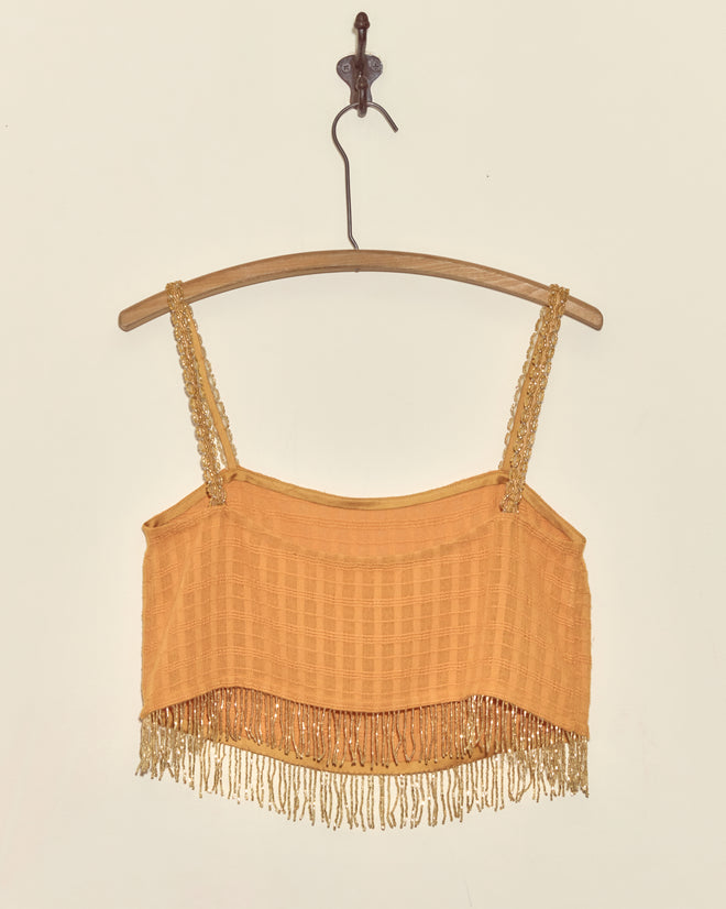 The Gilt Rosetta Tank - S is an orange cropped top with decorative straps, vintage beaded trim, a fringe hem, and hand-mended details for unique charm. Displayed on a wooden hanger against a plain wall.