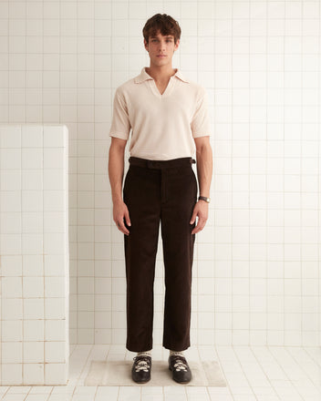 A man stands in a tiled room, wearing a beige shirt, Gingerbread Trousers in brown corduroy, and black shoes.