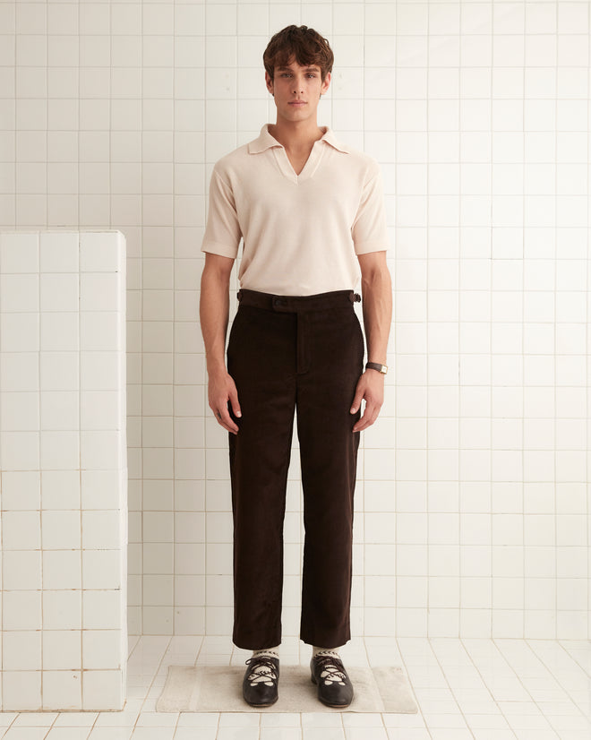 A man stands in a tiled room, wearing a beige shirt, Gingerbread Trousers in brown corduroy, and black shoes.