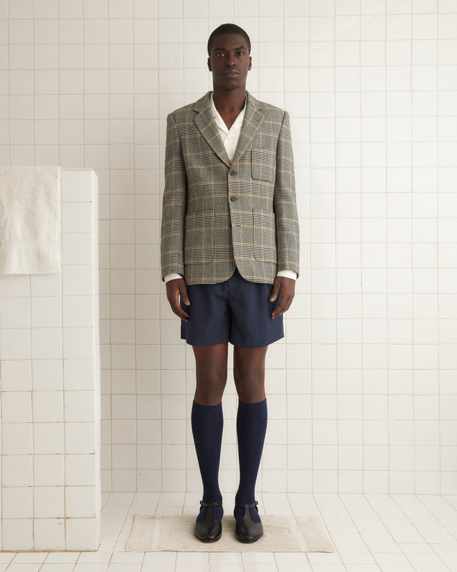 Man in a Golden Grid Suit Jacket - 40, white shirt, and navy shorts, with knee-high socks standing on a tiled floor in a minimalist room.