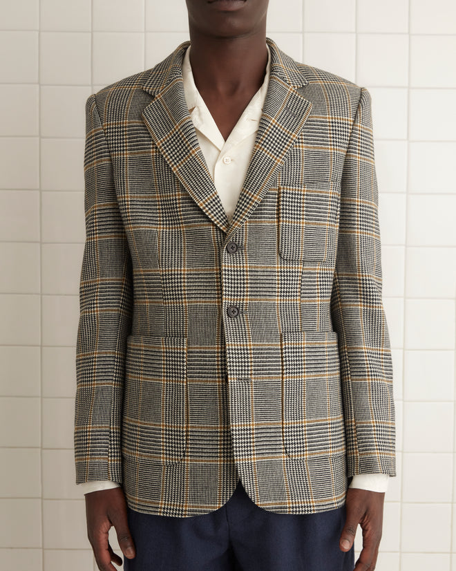 Man in a Golden Grid Suit Jacket - 40, white shirt, and navy shorts, with knee-high socks standing on a tiled floor in a minimalist room.