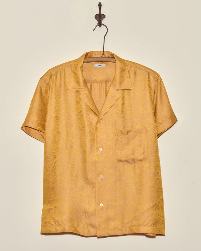 The Golden Shell Short Sleeve Shirt (Size S), crafted from golden-yellow deadstock jacquard, hangs on a metal hook against a plain wall.