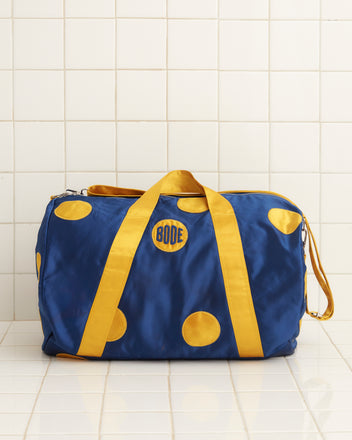 The Gold Medal Gym Bag, featuring a vibrant blue design with yellow dots and straps reminiscent of a jockey jacket, is showcased against a tiled wall.