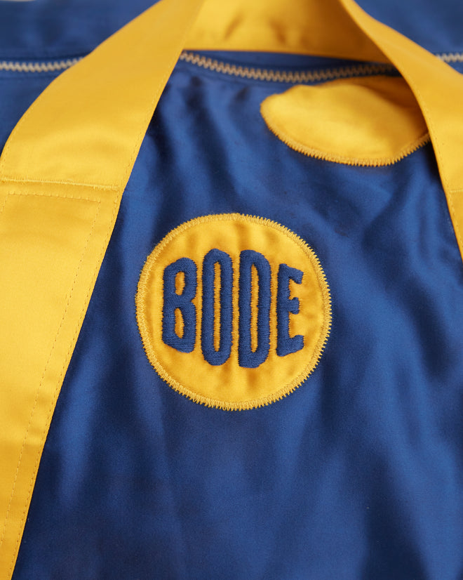 The Gold Medal Gym Bag, featuring yellow straps and charming polka dots on a blue background, reminiscent of a vibrant jockey jacket, sits stylishly on a tiled surface.