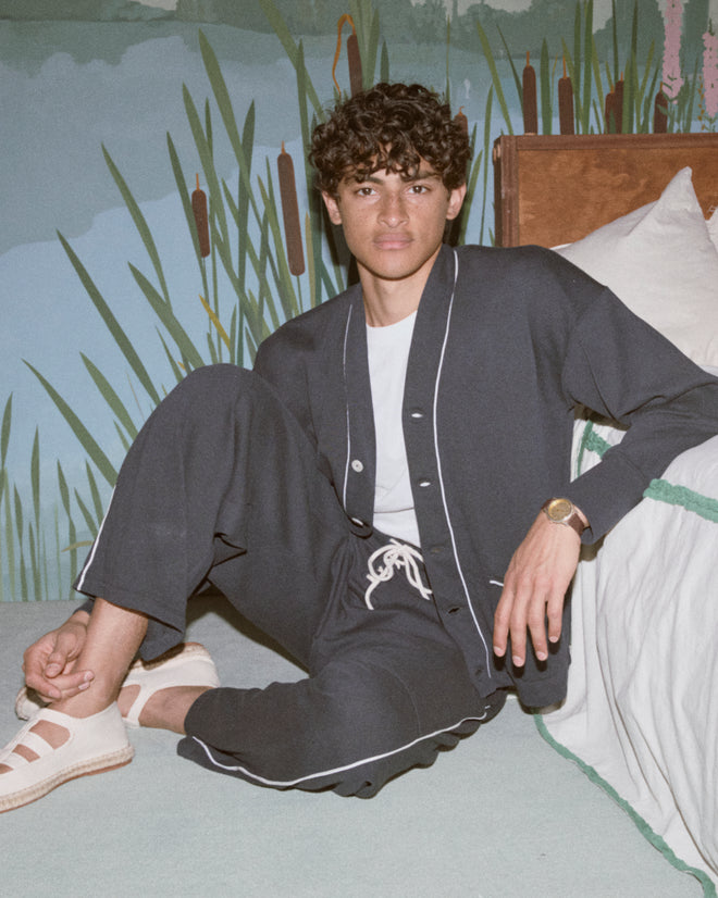 A young man in Lace-Front Sweatpants made of dark heavyweight fabric sits on the floor by a bed, leaning against a mural with tall green plants.