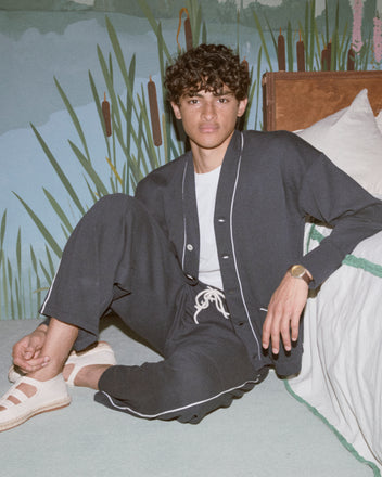 A young man sits on the floor by a bed against a nature-themed wall mural, wearing black Lace-Front Sweatpants with white trim.