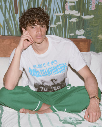 A young man with curly hair sits cross-legged on a bed, wearing green Sophomore Sweatpants and a heavyweight graphic T-shirt.