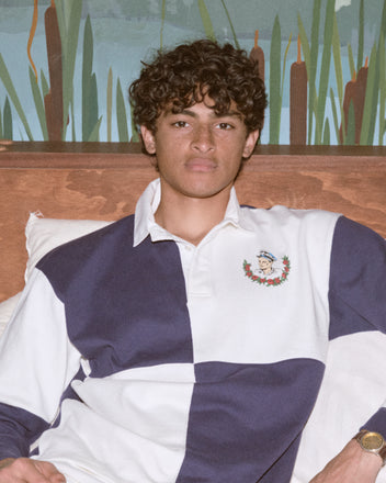 A young man with curly hair sits on a bed wearing the Grandfather Polo, a classic blue and white 1980s-inspired rugby shirt with a crest, made from 100% cotton.