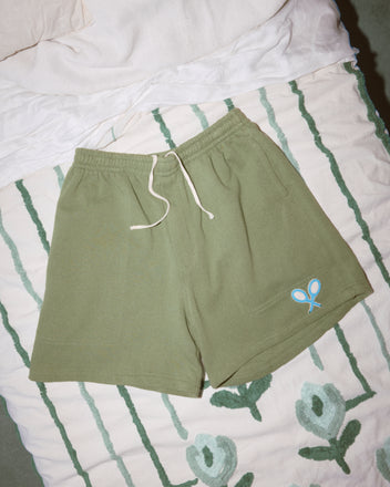 Beaded Racquet Patch Shorts in olive green with a drawstring, blue tennis racket logo, and novelty tennis patches, displayed on a patterned bedspread.