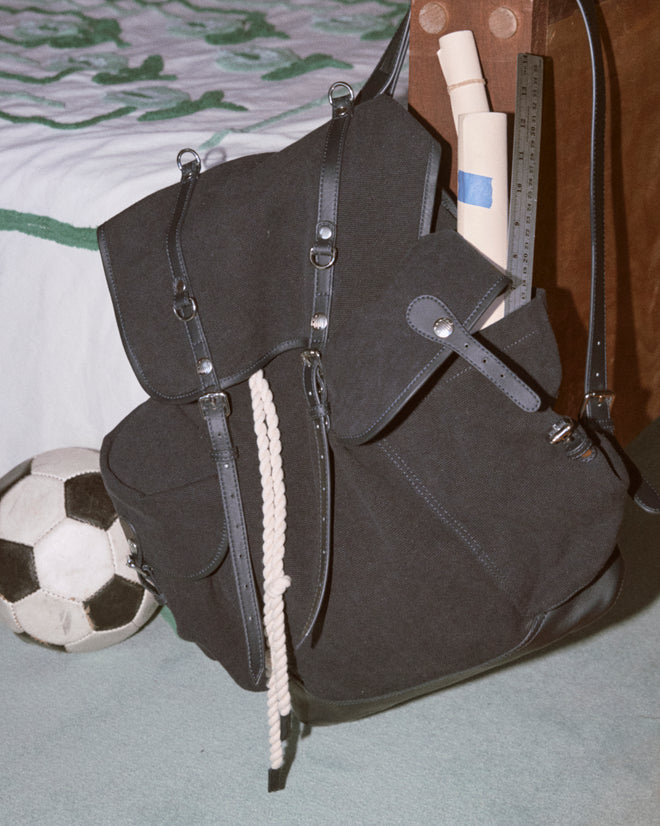 A Canvas Rucksack - Black, crafted from heavyweight canvas with leather accents, sits by a bed filled with a rope, rolled paper, and ruler, accompanied by a soccer ball.