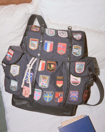 The Interrail Rucksack, a vintage-inspired black backpack adorned with colorful country patches, rests on a white cushion.