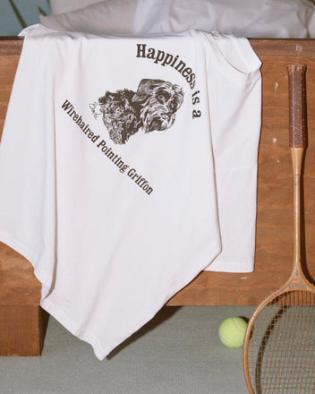 The Happiness Tee, a white 100% cotton shirt featuring a cute dog graphic and “Happiness is a Wirehaired Pointing Griffon,” rests on a wooden bench.