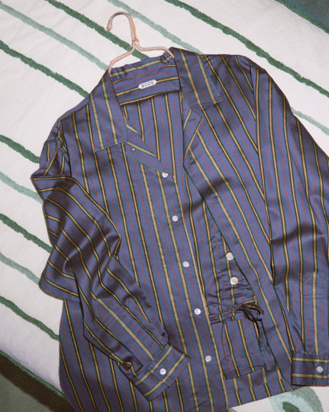 The Alumni Stripe Long Sleeve Shirt- Blue, a men's striped button-up made from a viscose silk blend, hangs neatly on a hanger and is displayed on a green and white striped blanket.