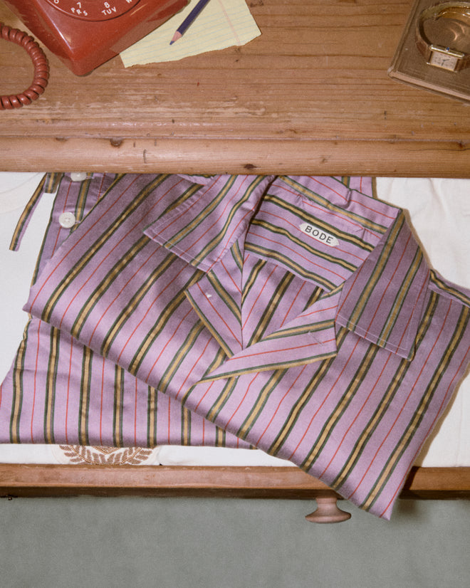 The Alumni Stripe Long Sleeve Shirt - Purple, featuring classic stripes and "BODE" labeling, sits folded in an open drawer beneath a wooden desk with a red rotary phone, evoking the charm of 1960s-1980s men's shirts.