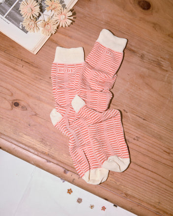 A pair of Micro Check Socks in Peach/Cream, inspired by vintage children's socks, displayed on a wooden table near flowers and a book.