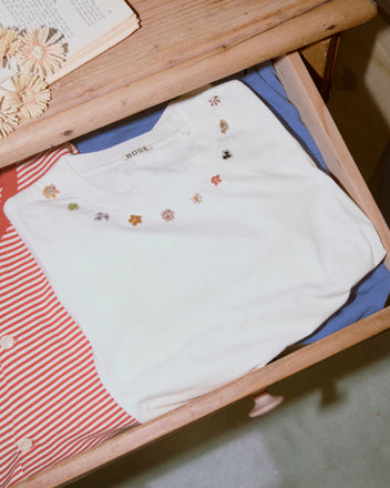 The Beaded Floral Necklace Tee, a white 100% cotton T-shirt with small embroidered flowers, is folded in a wooden drawer next to other colorful clothes.