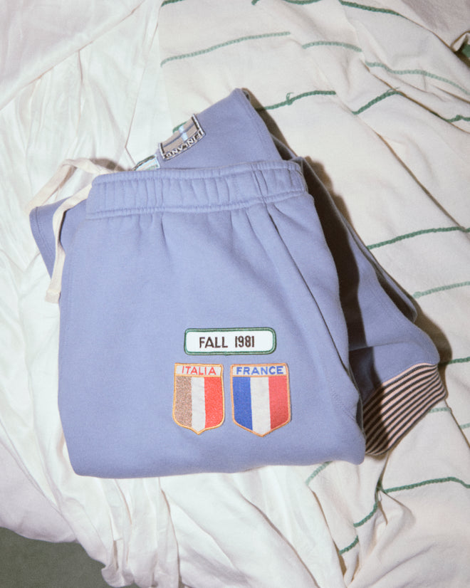 Warmup Patch Joggers in light blue, featuring vintage "FALL 1981," Italia, and France patches with striped cuffs, are displayed on top of a white striped shirt.