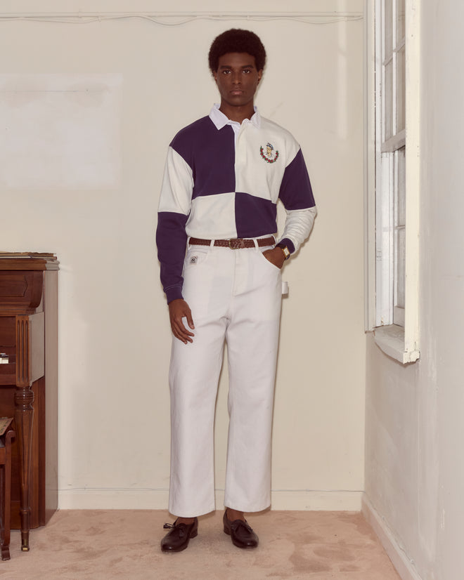 A man wears the Grandfather Polo paired with white cotton trousers, channeling 1980s style as he stands indoors by a window and a piano.
