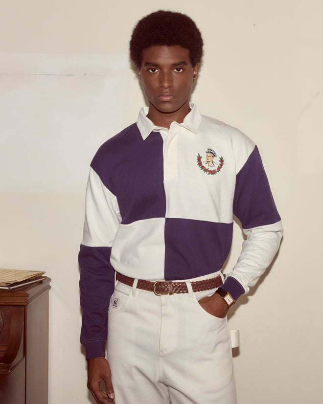 A person wearing the Grandfather Polo, a blue and white cotton rugby shirt with an 1980s-inspired embroidered crest, stands indoors by a wooden desk.