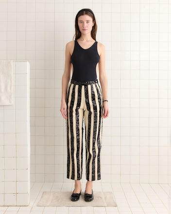 In a tiled room, a person wears a black tank top and Grandstand Hall Trousers. The high-rise fit complements their stance as sequin stripes catch the light, adding sparkle to the ensemble while their hands rest by their sides.