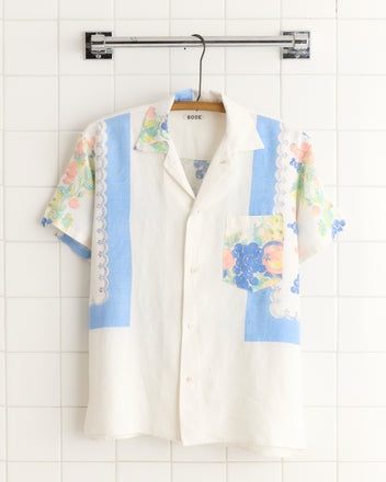 The Grapevine Short Sleeve Shirt (XS/S) features blue and floral patchwork, vintage tablecloth-inspired, displayed on a wooden hanger against white tiles.