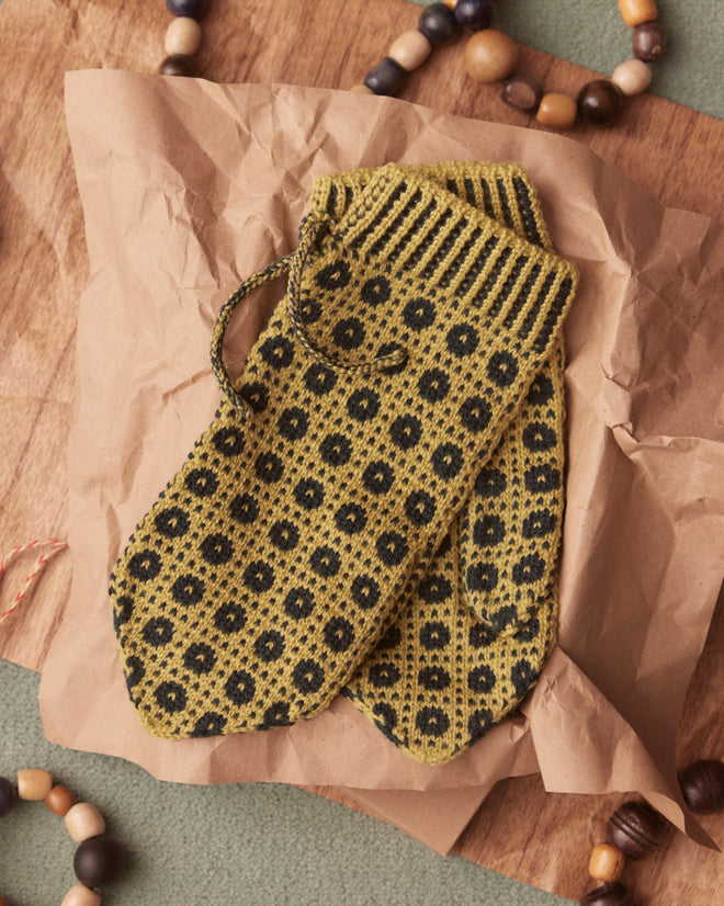 The Geo Knit Mittens in a green color, crafted from merino wool, rest on crumpled brown paper, surrounded by wooden beads.