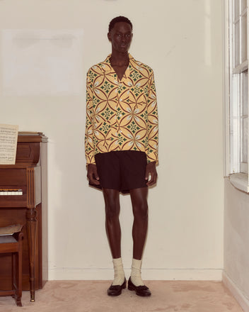 A person stands indoors beside a piano, wearing the Greer Long Sleeve Shirt with a geometric pattern, paired with black shorts, socks, and loafers.