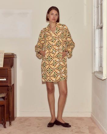 A woman wearing the Greer Quincy Dress, featuring a 1960s-inspired geometric pattern, stands indoors by a window and piano with her hands in her pockets, looking at the camera.