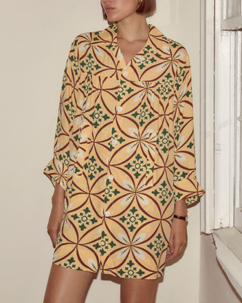 A woman stands near a window wearing the Greer Quincy Dress, a 1960s-inspired yellow dress featuring geometric and floral patterns on soft viscose fabric.