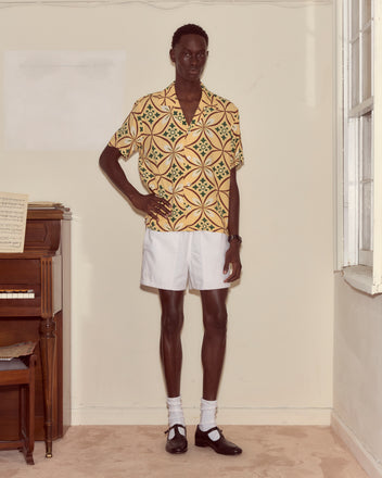 A man in a Greer Short Sleeve Shirt with geometric patterns, white shorts, and black shoes stands indoors next to a piano and a window.