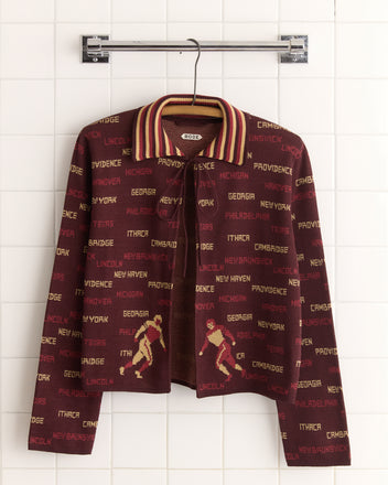 The Gridiron Cardigan, a brown jacket with city names and skiers embroidered like a 1940s sweater, hangs on a metal rod against a tiled wall.