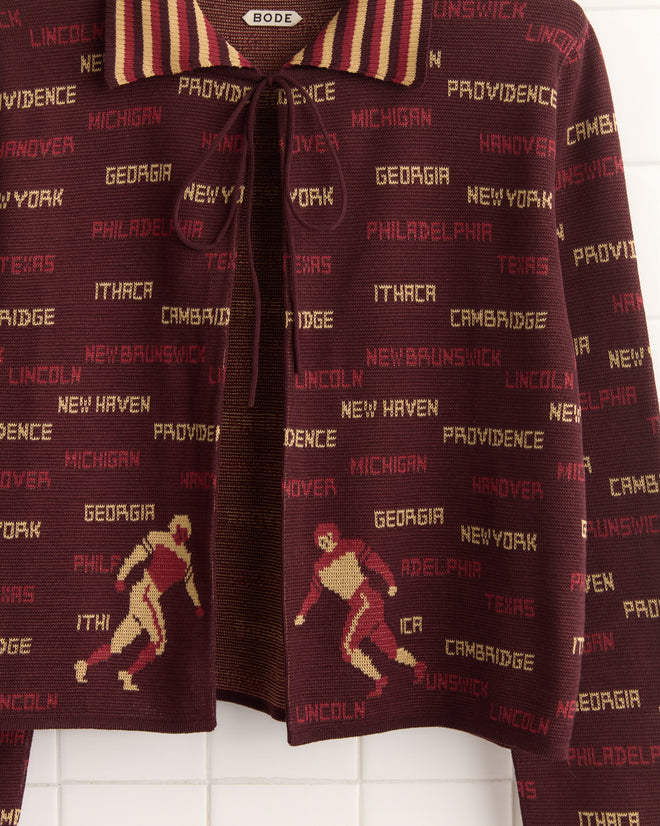 The Gridiron Cardigan, a maroon 1940s-style sweater adorned with city names like Providence and New York and skier graphics, hangs against a white backdrop.