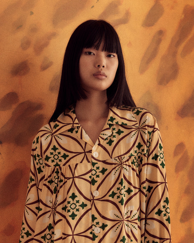 A person with long, dark hair wearing the Greer Quincy Dress, inspired by the 1960s style, stands against a warm-toned, abstract background.