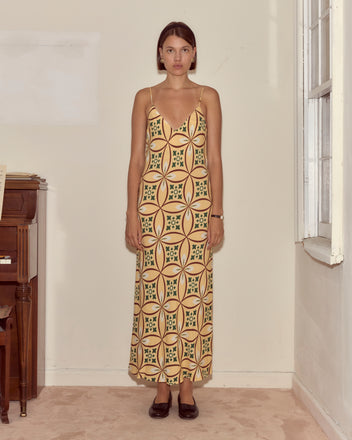 A woman wearing the Greer Slip Dress stands indoors by a piano and window, looking straight at the camera.