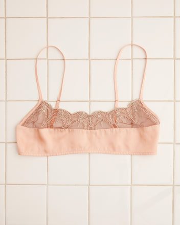 The Half Shell Bra, featuring a light pink lace design with thin straps and mother-of-pearl buttons, is laid flat on a tile surface.