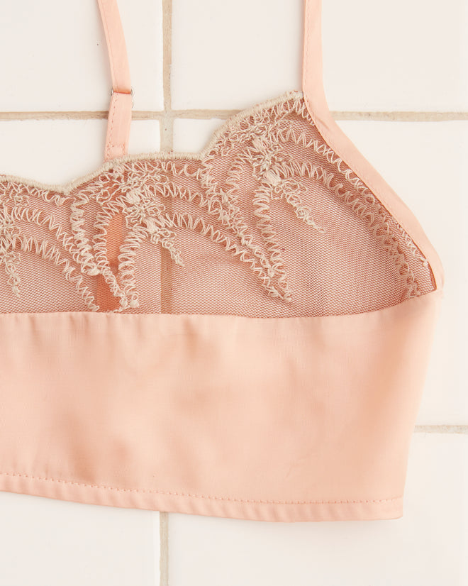 The Half Shell Bra, featuring a light pink lace design with thin straps and mother-of-pearl buttons, is laid flat on a tile surface.