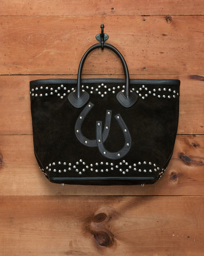 The Studded Horseshoe Tote, crafted from black cow suede with studded patterns and black handles, is accented by a leather horseshoe appliqué and hangs on a hook against a wooden wall.