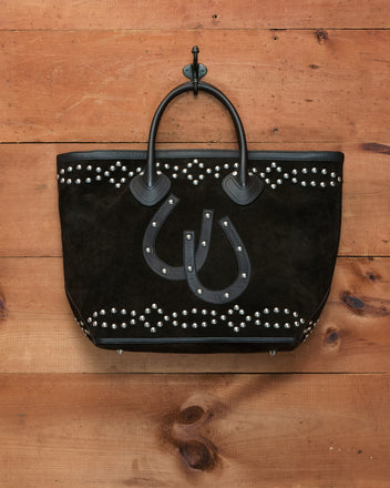 The Studded Horseshoe Tote, crafted from black cow suede with studded patterns and black handles, is accented by a leather horseshoe appliqué and hangs on a hook against a wooden wall.