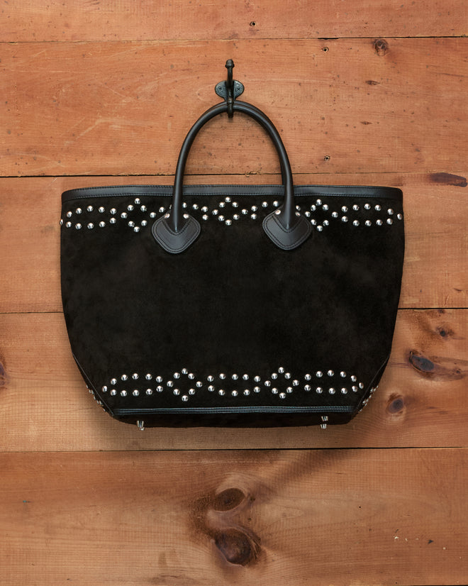 The Studded Horseshoe Tote, crafted from black cow suede and featuring silver studs with a leather horseshoe appliqué, hangs on a hook against a wooden wall.